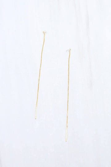 A pair of delicate gold drop earrings with thin chains and small cylindrical dangling charms, displayed against a soft white fabric background.