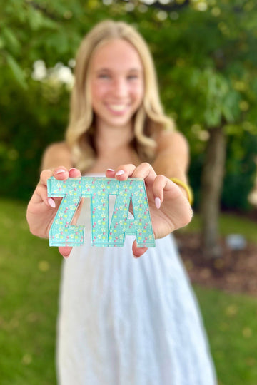 Woman holding 'ZITA' letters outdoors with greenery in the background