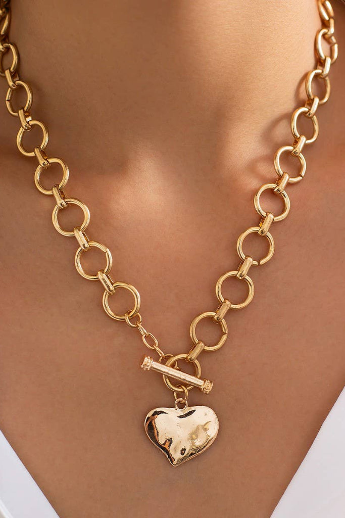 A close-up of a gold chain necklace with a toggle clasp and a polished heart-shaped pendant, worn on a model's neck over a white collared shirt.