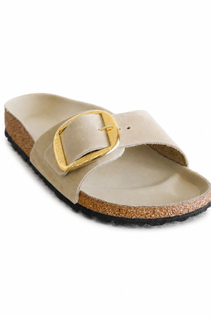 A single beige slide sandal with a wide strap featuring a large gold buckle, a cork midsole, and a black outsole, shown on a plain white background.
