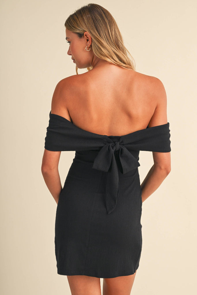 A woman shown from the back wearing a black off-the-shoulder mini dress with a large bow detail at the back, against a plain beige studio background.