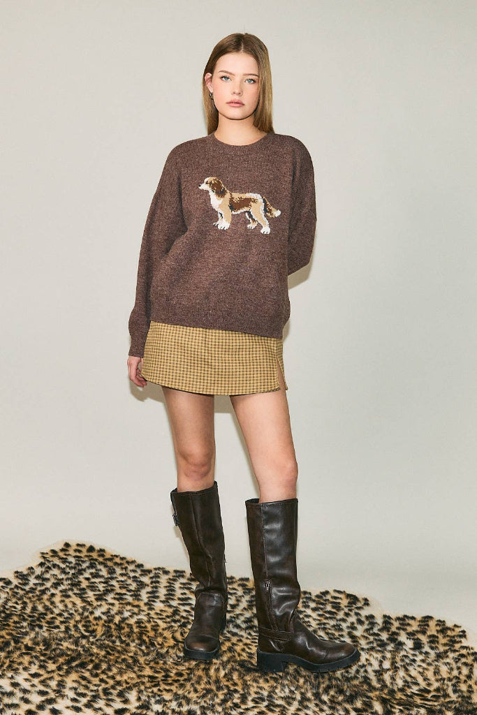 A model stands on a leopard-print rug wearing a brown knit sweater with a dog motif, a beige mini skirt, and black knee-high boots against a plain light gray studio background.