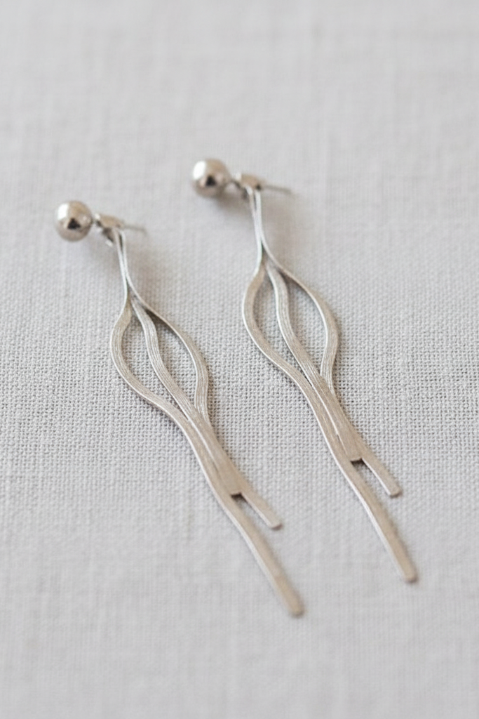 A pair of silver drop earrings with a minimalist, abstract leaf-like design and small spherical studs, displayed on a textured light beige linen fabric background.