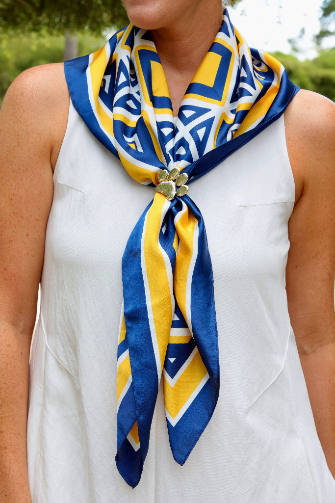 A close-up of a woman wearing a white sleeveless top and a blue and yellow patterned silk scarf tied at the collar with a decorative gold-toned knot.