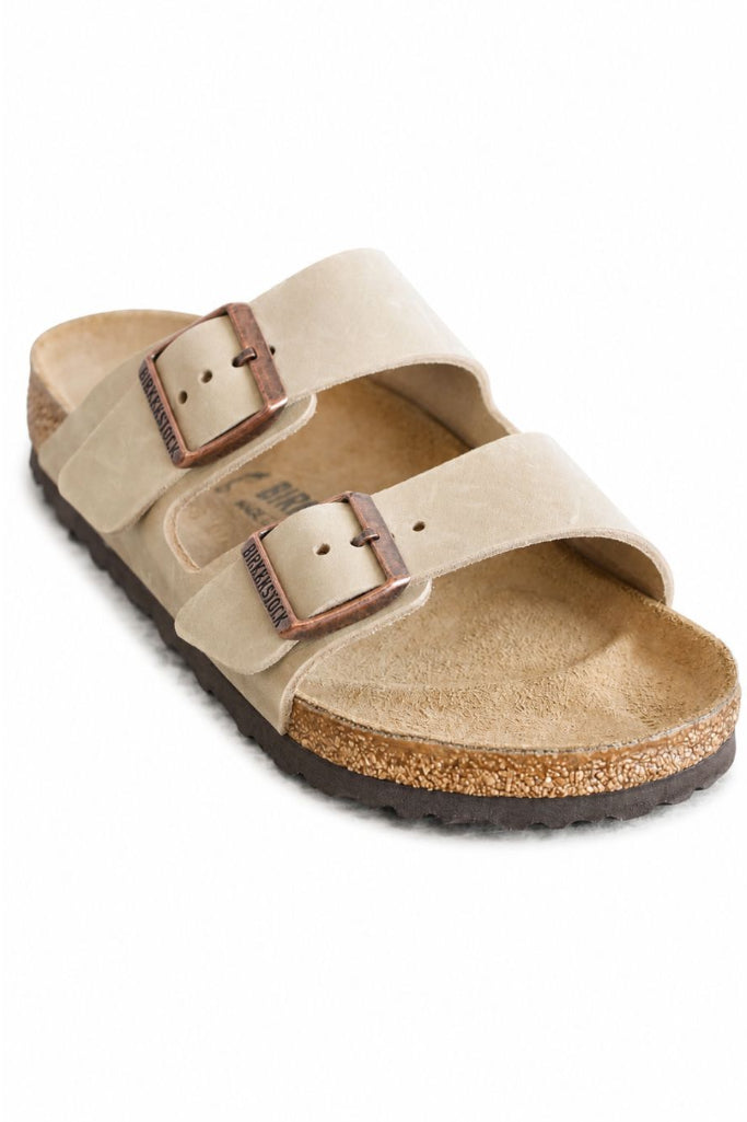 A single beige double-strap sandal with adjustable metal buckles, a tan footbed, and a cork-look midsole on a white background.