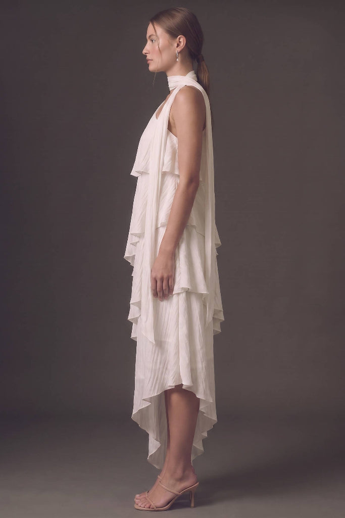 A woman in profile wearing a sleeveless, high-neck, tiered white dress with a high-low hem, paired with nude high-heeled sandals, set against a dark gray studio background.