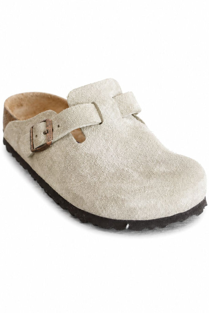 A light beige suede clog shoe with a double strap closure and a brown buckle, shown from a three-quarters angle on a white background.