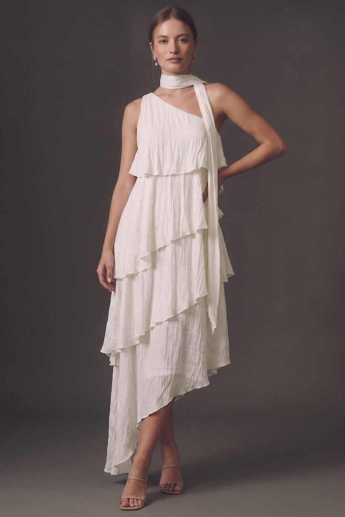 A woman models a white one-shoulder ruffled dress with a matching neck scarf and nude high-heeled sandals against a dark gray studio background.