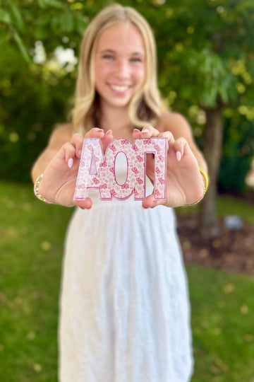 A smiling young woman in a white dress stands outdoors in a garden, holding up decorative pink letters spelling 'AOT' with a floral pattern.
