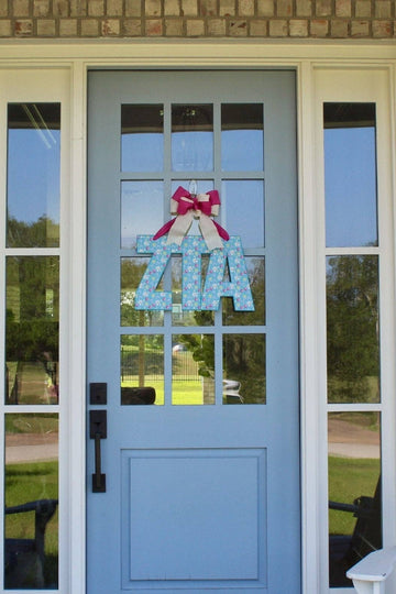 A light blue front door with glass panes, decorated with a blue patterned 'ZIA' letter sign and a red bow. The door is set in a beige brick wall with two black lantern-style wall lights and white Adirondack chairs with blue striped cushions on either side.