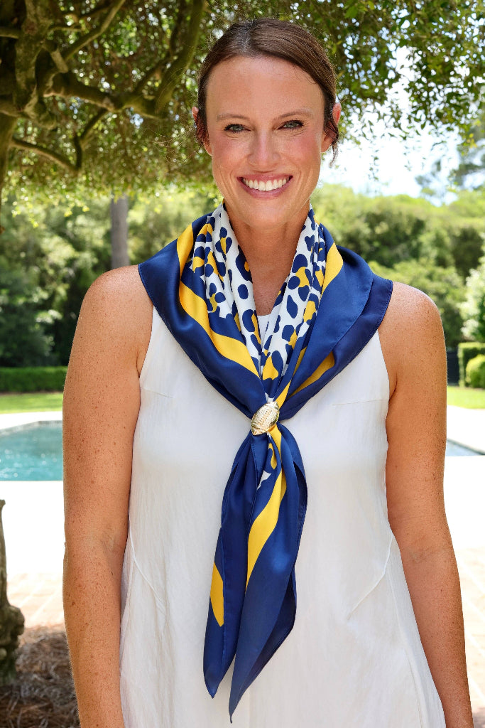 A woman outdoors wearing a white sleeveless dress and a navy blue silk scarf with yellow and white patterns, tied around her neck.