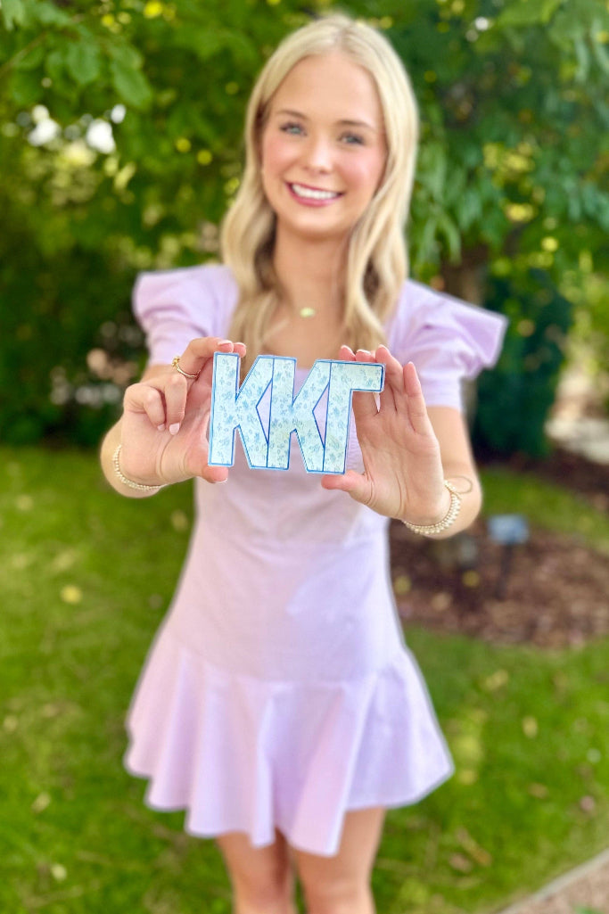 A smiling blonde woman outdoors in a light lavender dress holds up a decorative square sign featuring the Greek letters K, K, and G in light blue with white marbled patterns.