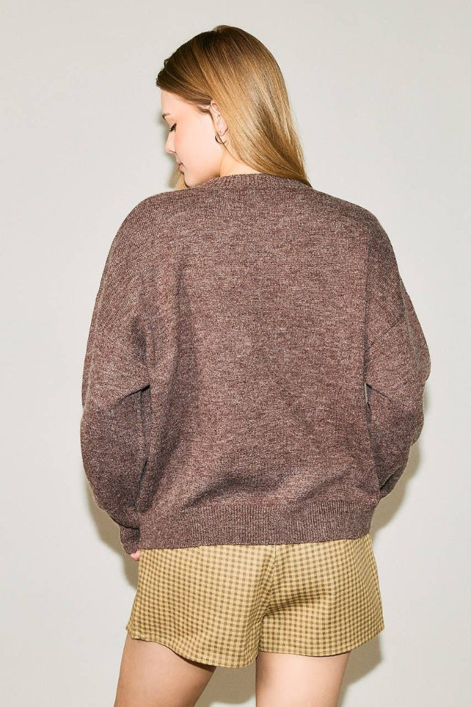 A woman shown from the back wearing an oversized heathered brown knit sweater and tan-and-black checkered shorts against a plain light gray background.