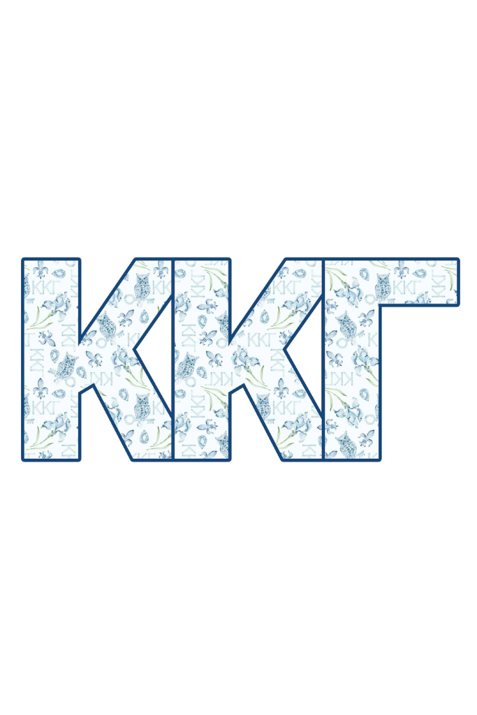 A smiling blonde woman outdoors in a light lavender dress holds up a decorative square sign featuring the Greek letters K, K, and G in light blue with white marbled patterns.