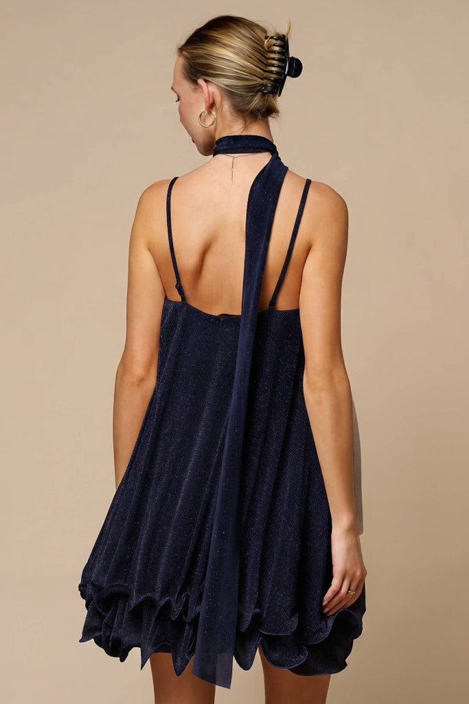 A woman shown from the back wearing a navy blue halter-neck dress with thin straps, a high open back, and a ruffled hem, set against a solid beige background.