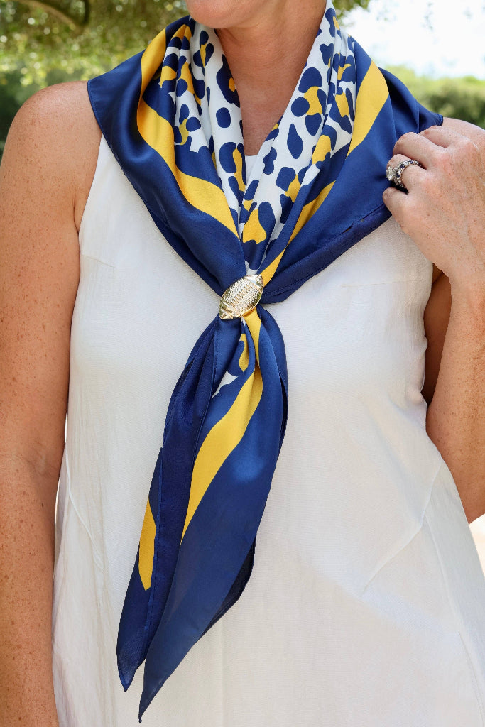 A woman wearing a navy blue and yellow patterned silk scarf tied around her neck with a decorative gold-toned clasp, paired with a white sleeveless top in an outdoor setting.