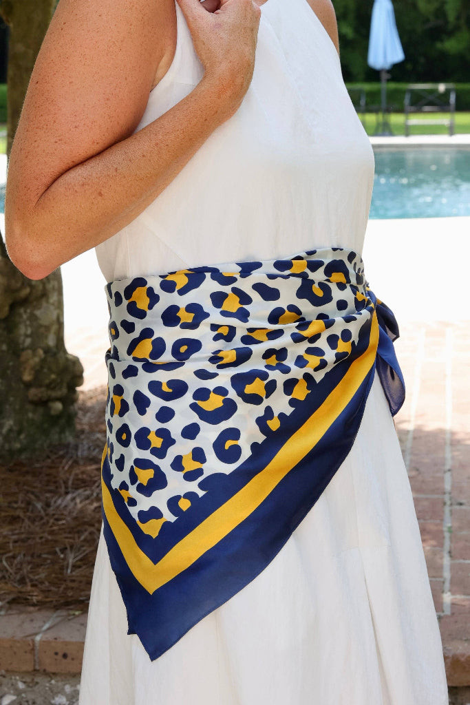 A navy blue and yellow leopard print scarf tied around the waist of a model wearing a white sleeveless dress, set outdoors next to a brick patio and swimming pool.