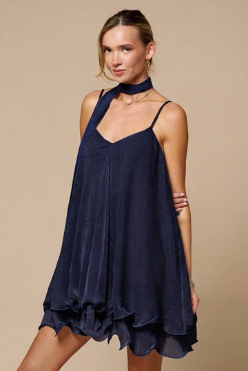 A woman modeling a navy blue halter-neck mini dress with a ruffled hem against a solid beige studio background.