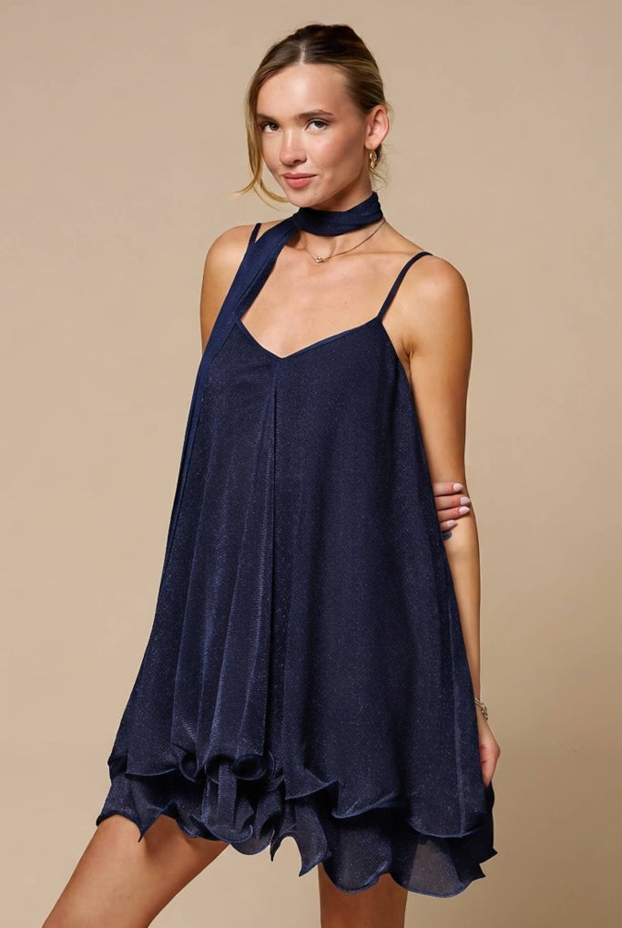 A woman modeling a navy blue halter-neck mini dress with a ruffled hem against a solid beige studio background.