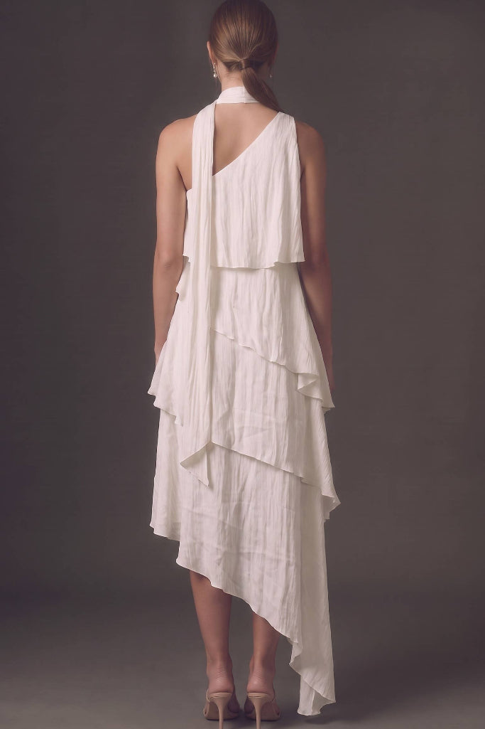 A woman viewed from the back wearing a white one-shoulder draped dress with layered ruffles and an asymmetrical hem, paired with nude high heels, against a dark gray studio background.